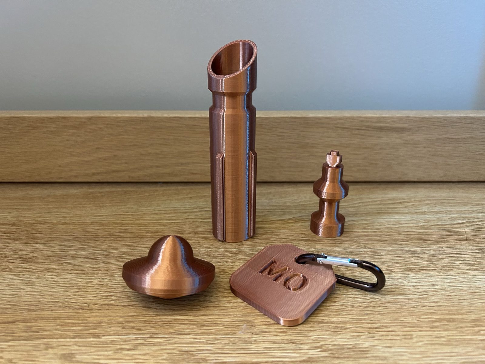 Four prints in copper filament: a telescoping lightsaber hilt, a chess piece, a UFO spinning top, and a bag tag stamped MO on a carabiner.